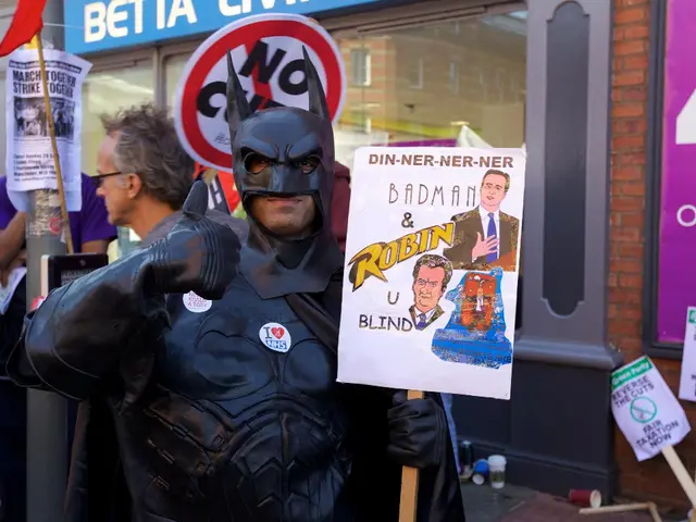 In the foreground of this image, there is a man in batman costume and holding a placard. On the...
