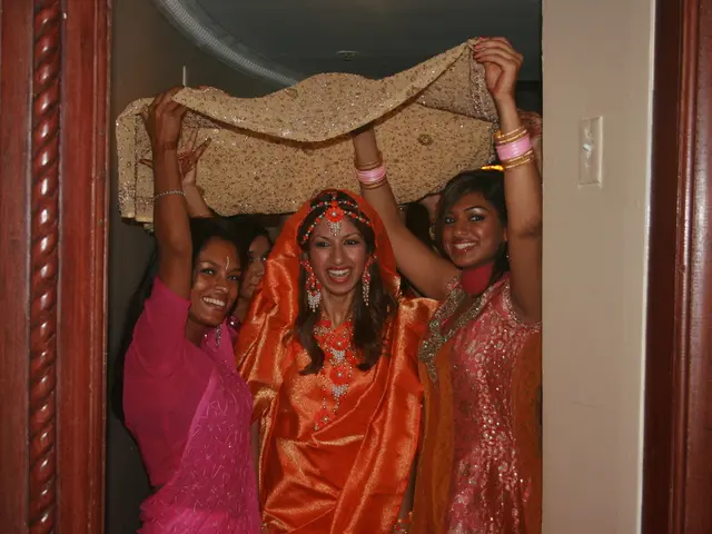 In the image there are few women standing and holding a cloth above under a bride groom in orange...